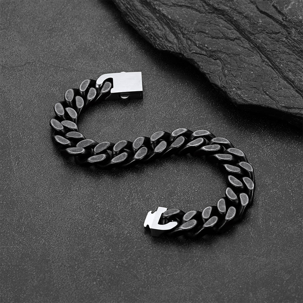 A black Cuban link chain bracelet, arranged in an S-shape on a dark textured surface with a slate prop.