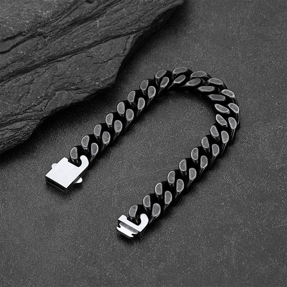 A black Cuban link chain bracelet with polished silver-toned metal ends, arranged in a U-shape on a dark textured surface with a slate prop.