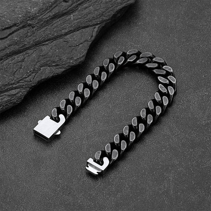 A black Cuban link chain bracelet with polished silver-toned metal ends, arranged in a U-shape on a dark textured surface with a slate prop.