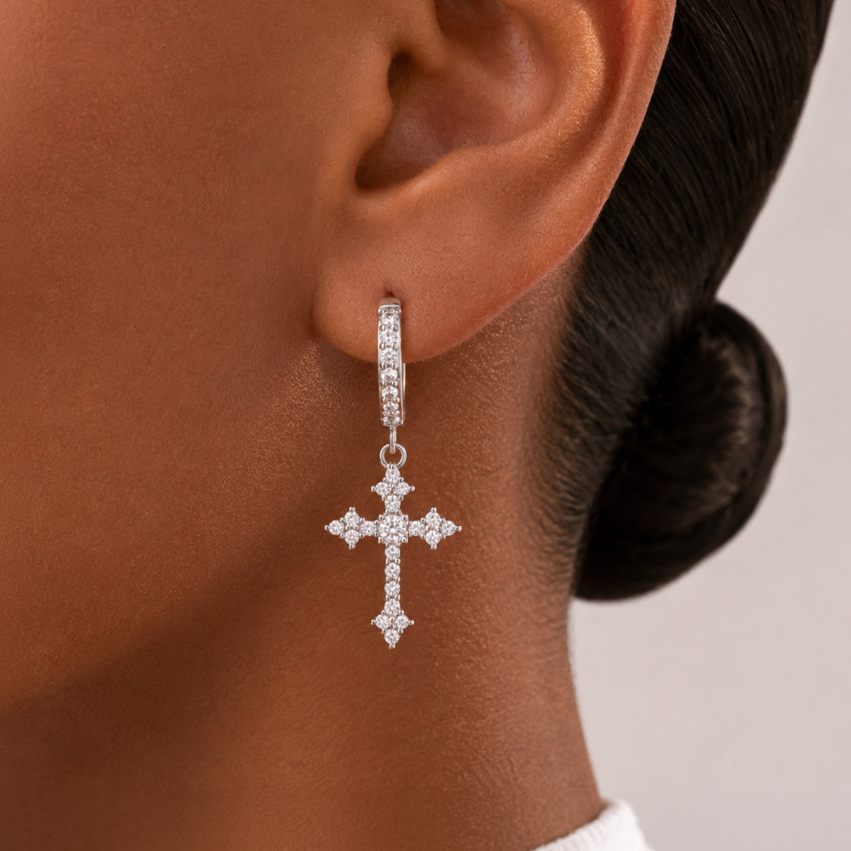 A close-up of a model's ear wearing a silver cross drop earring adorned with small clear stones.