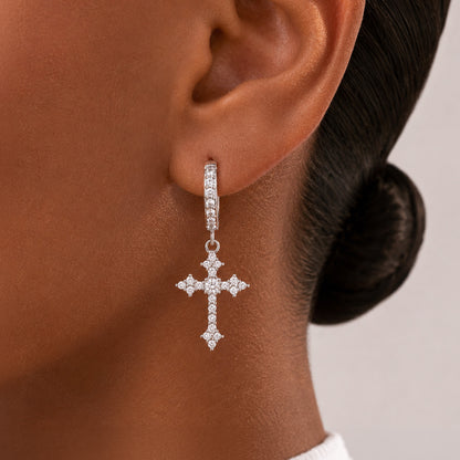 A close-up of a model's ear wearing a silver cross drop earring adorned with small clear stones.