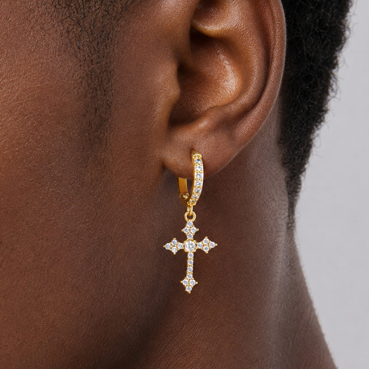 A close-up of a person's ear wearing a gold huggie hoop earring with a dangling cross with small clear stones.