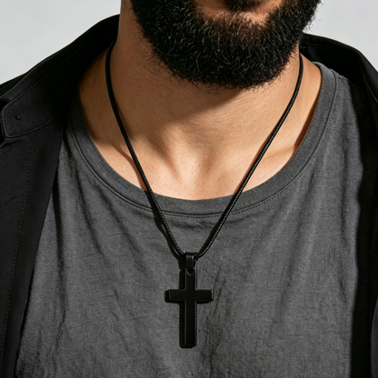 A close-up of a man's neck and chest wearing a matte black cross pendant necklace over a dark gray t-shirt and black jacket.