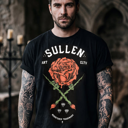 A man with tattooed arms wearing a black graphic t-shirt featuring a large red rose and crossed green stems with the text 'SULLEN' and 'ART CLTV'. The background is a dark, moody stone interior with blurred candles.