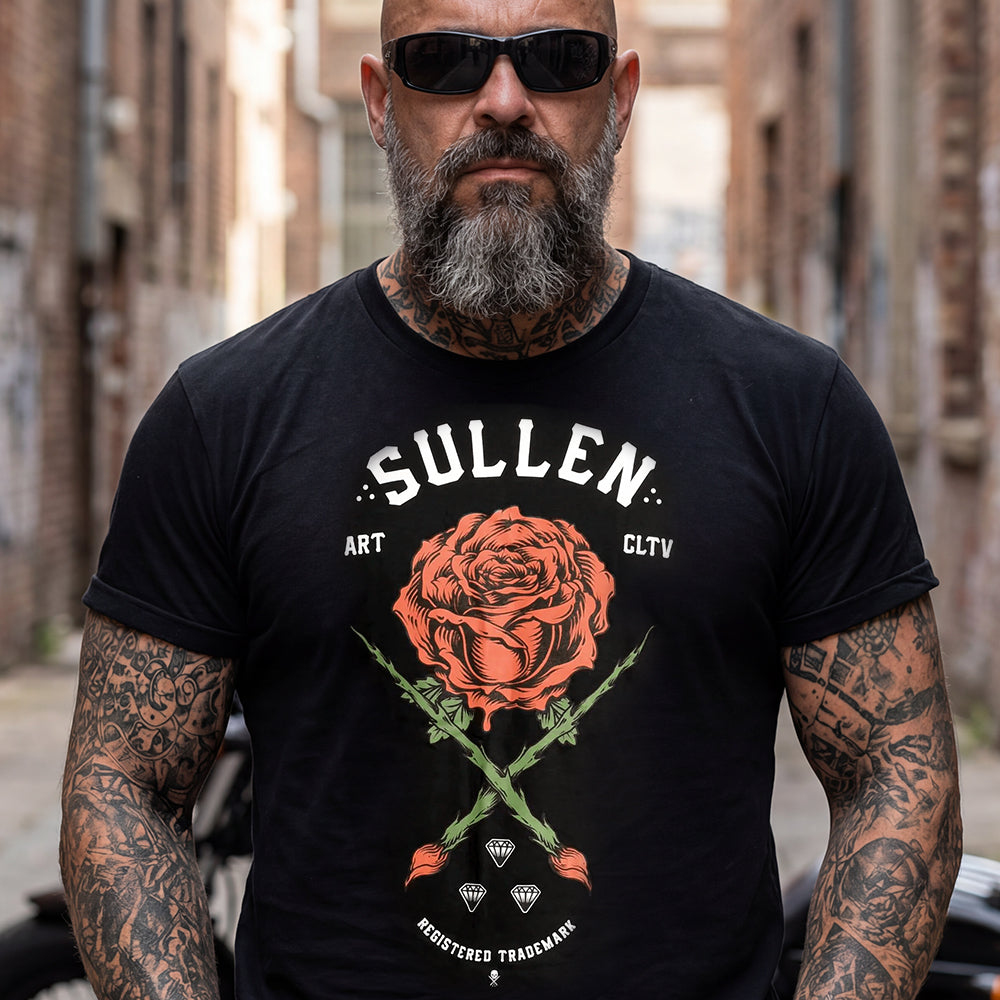 A tattooed man wearing a black graphic t-shirt with a large red rose and the text 'SULLEN ART CLTV' on the front, standing in a blurred urban alleyway.