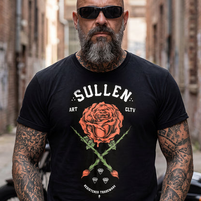 A tattooed man wearing a black graphic t-shirt with a large red rose and the text 'SULLEN ART CLTV' on the front, standing in a blurred urban alleyway.