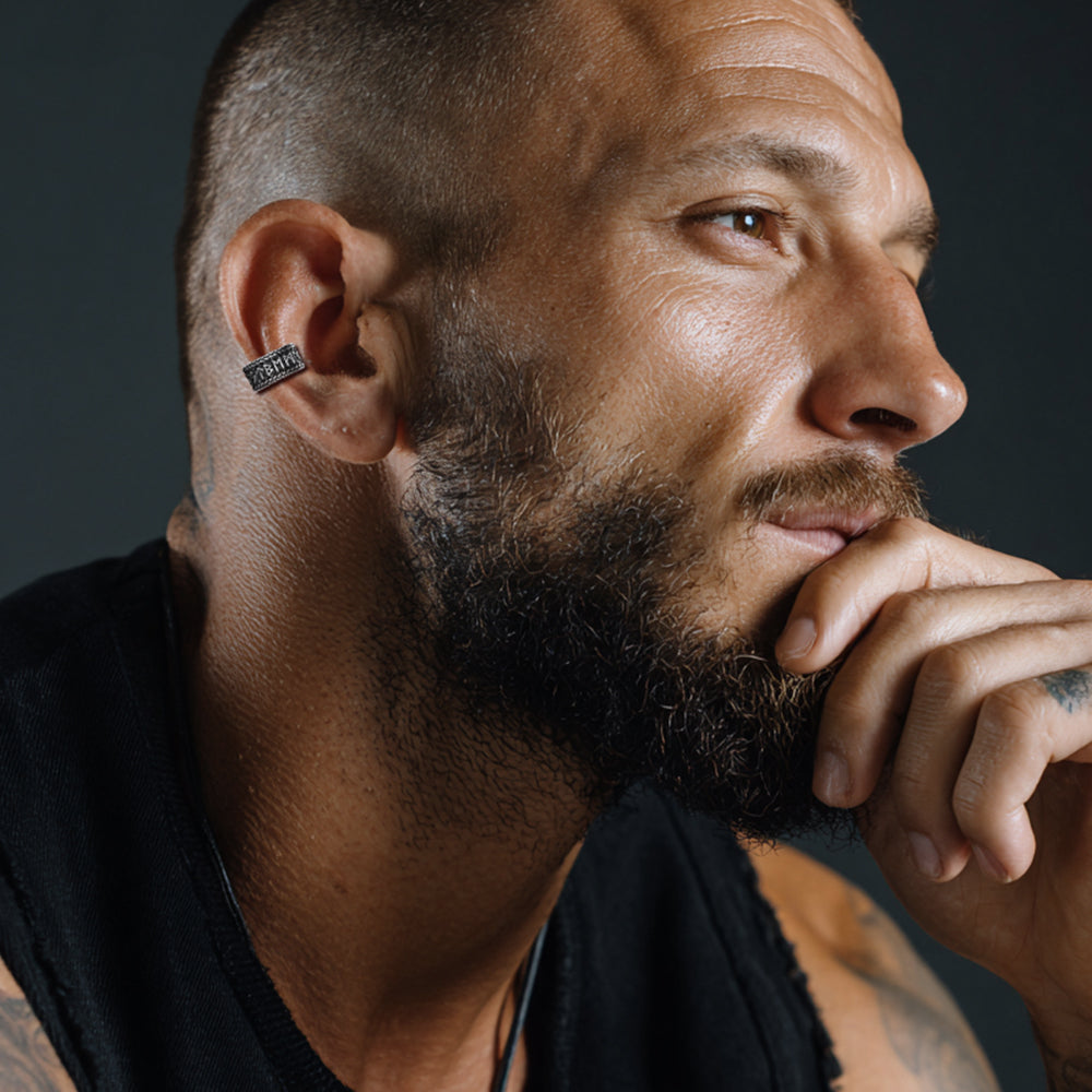 Man with a beard and tattoos on his arm, wearing a black shirt, against a dark background.