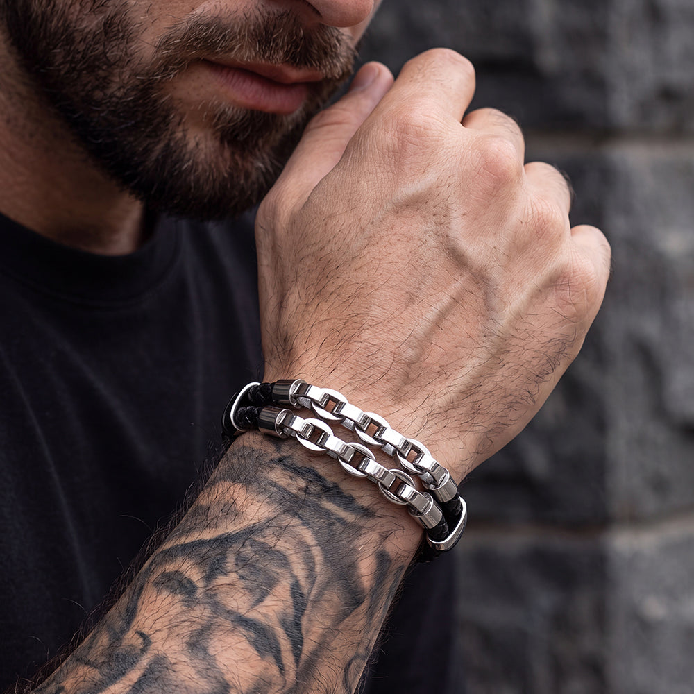 A close-up of a man's tattooed forearm and hand wearing two interlocking metallic link bracelets