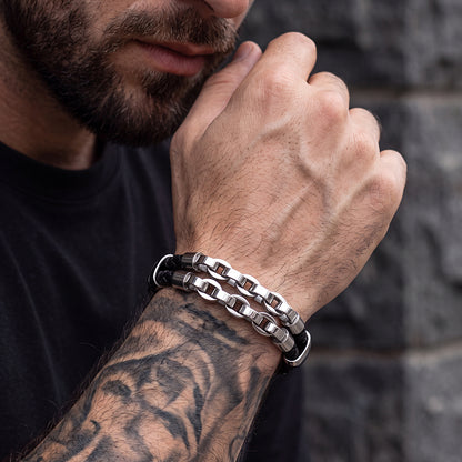 A close-up of a man's tattooed forearm and hand wearing two interlocking metallic link bracelets