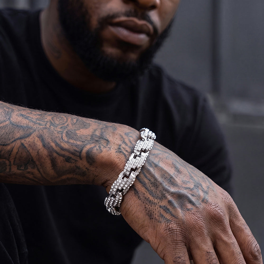 A close-up of a man's tattooed arm and hand wearing a chunky silver chain bracelet paved with clear stones.