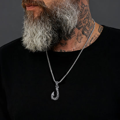 Man wearing a black shirt with a silver necklace featuring a hook pendant against a dark background