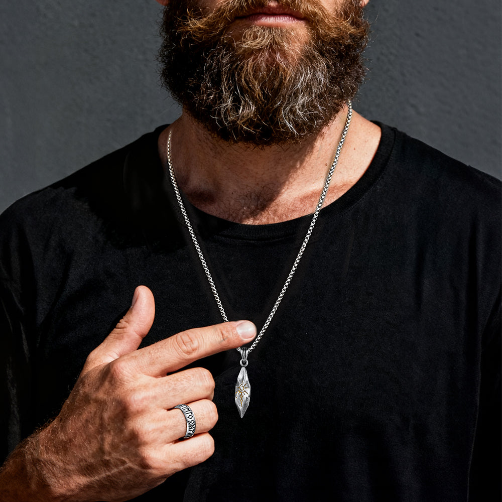 Man wearing a black shirt and a silver necklace with a pendant, pointing to the necklace.