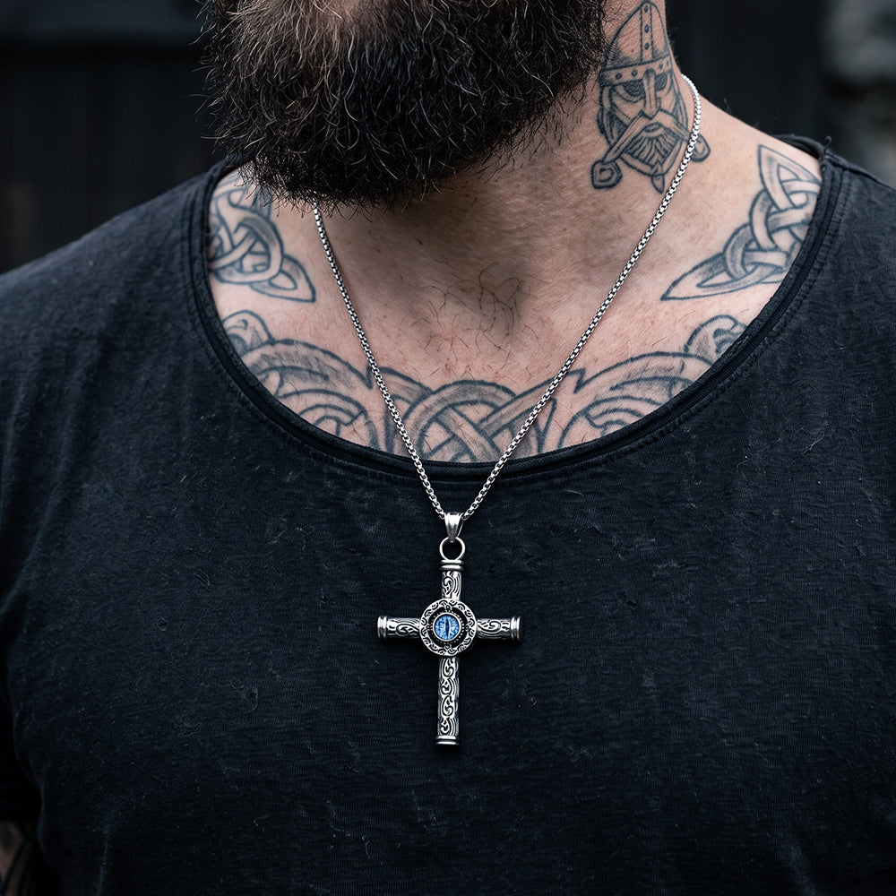 A close-up of a man's neck and chest wearing a silver-toned cross pendant necklace with a blue stone in the center, over a black t-shirt.