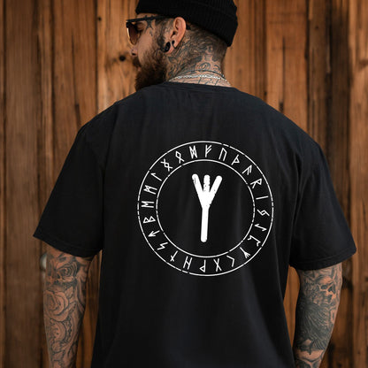 Person wearing a black t-shirt with a white symbol and text on the back, standing against a wooden wall.
