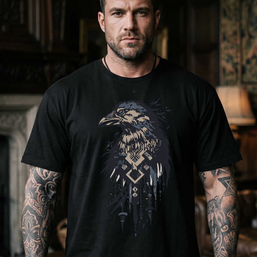 A man wearing a black graphic t-shirt with a detailed raven design in shades of blue, gray, and beige, set against a dark, atmospheric indoor background.
