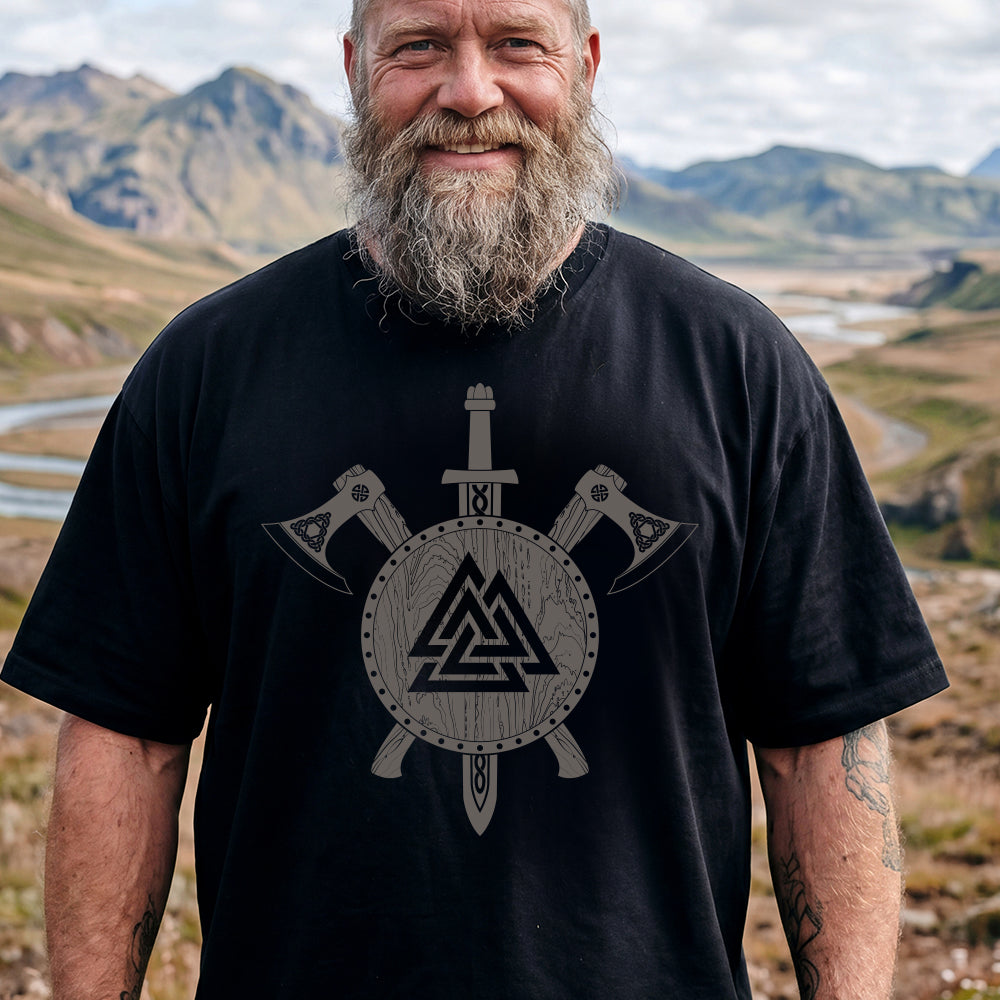 Man wearing a black Viking Valknut Shield Sword & Axes T-Shirt, standing in a mountainous landscape.