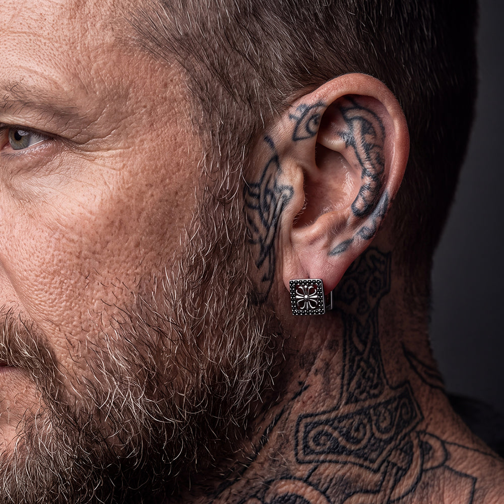 Close-up of a man's ear wearing a square silver stud earring with a cross and small black stones.